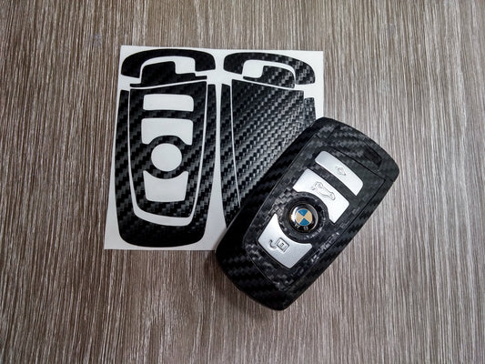 Carbon key wrap kit pieces on the backing sheet beside a wrapped BMW blade fob. Compatible with BMW, not genuine BMW parts.