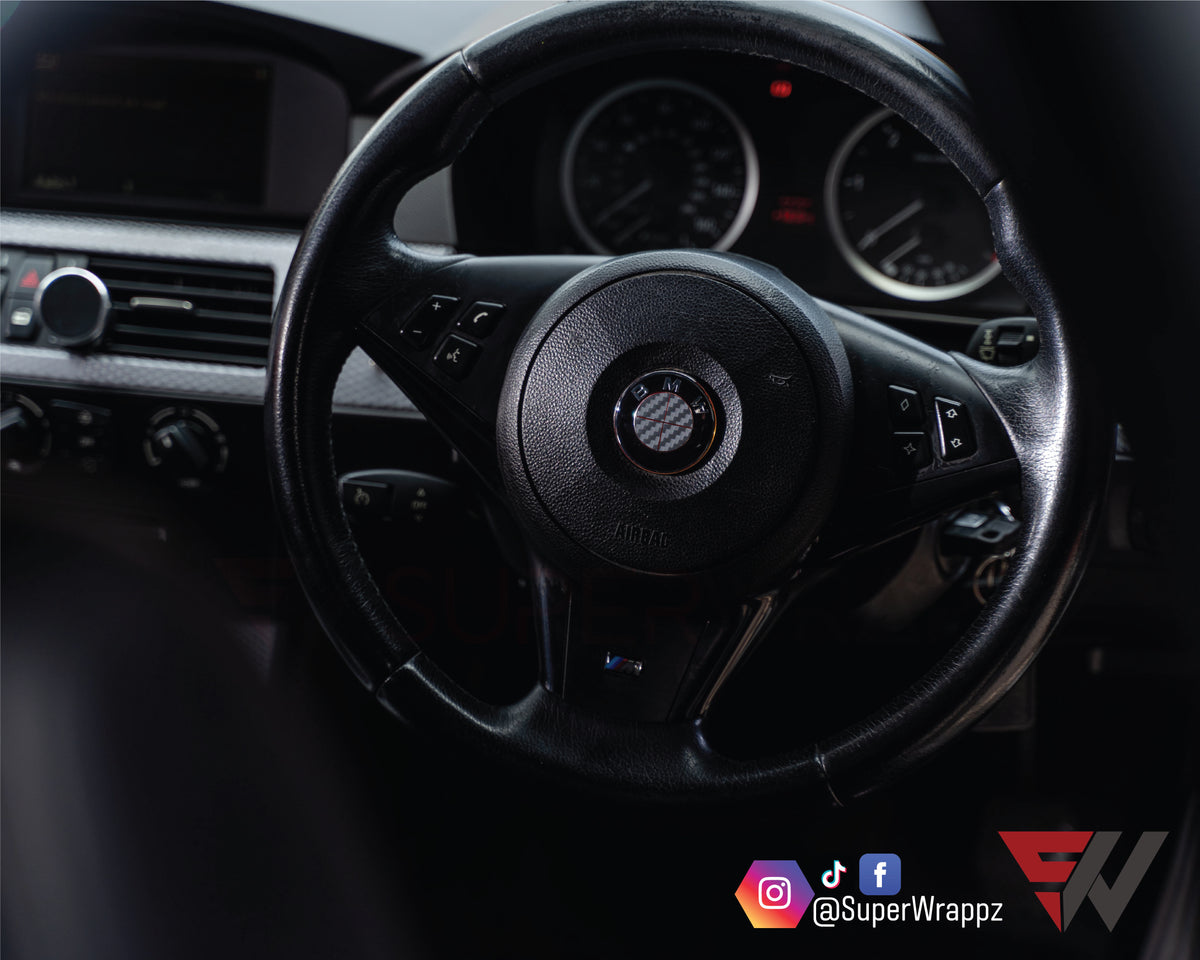 Full Dark Grey Carbon BMW badge overlay applied to a steering wheel centre badge, close-up